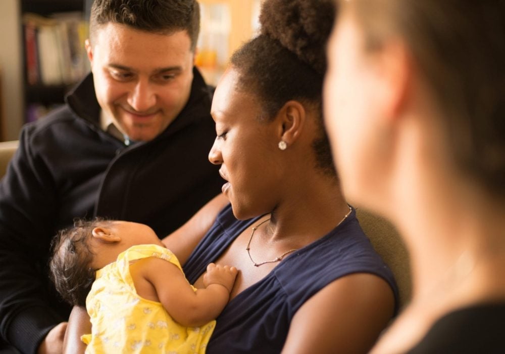 Woman holding an infant while a smiling man sitting beside her looks on.