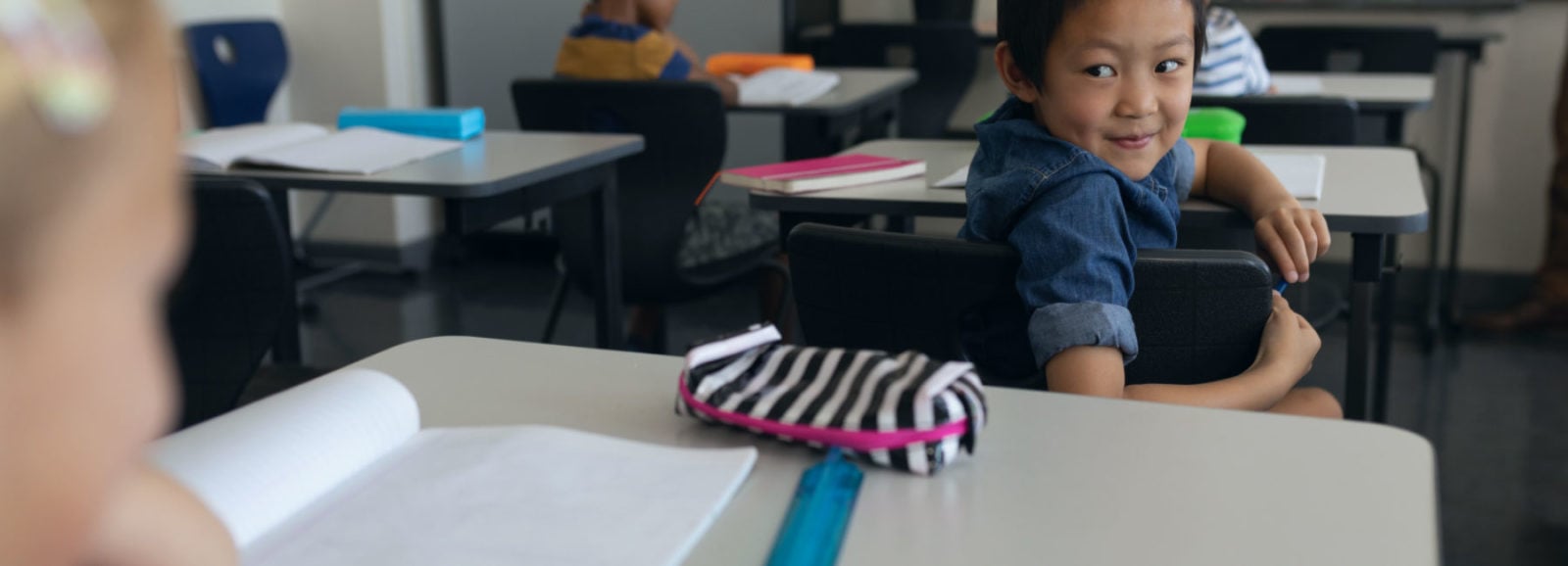 Smiling young boy in classroom looking back at another student behind him.