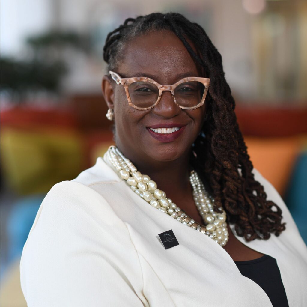 Woman with medium length braided hair wearing a white blazer, pearl necklace and glasses smiling at the camera