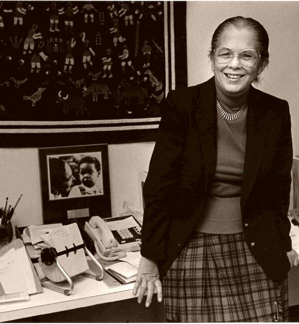 black and white image of Barbara Taylor Bowman leaning against a desk wearing a black blazer and plaid skirt