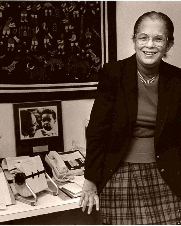 black and white image of Barbara Taylor Bowman leaning against a desk wearing a black blazer and plaid skirt
