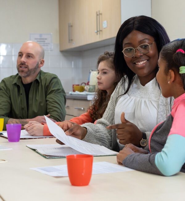 A medium group of children sit around a table with two adult therapists in a community centre in Whitley Bay, North East England. The diverse group of children are engaged in a supportive counselling session, with some looking attentive and others actively participating in the discussion. The therapists are warm and approachable, creating a nurturing environment. The room is brightly lit and welcoming, with comfortable seating, reflecting a safe space for sharing and healing. Videos are also available similar to this scenario.