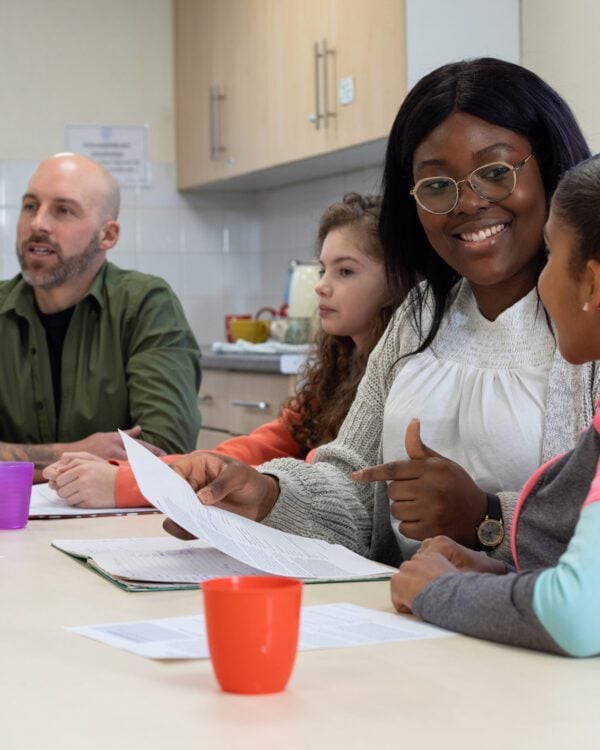 A medium group of children sit around a table with two adult therapists in a community centre in Whitley Bay, North East England. The diverse group of children are engaged in a supportive counselling session, with some looking attentive and others actively participating in the discussion. The therapists are warm and approachable, creating a nurturing environment. The room is brightly lit and welcoming, with comfortable seating, reflecting a safe space for sharing and healing. Videos are also available similar to this scenario.