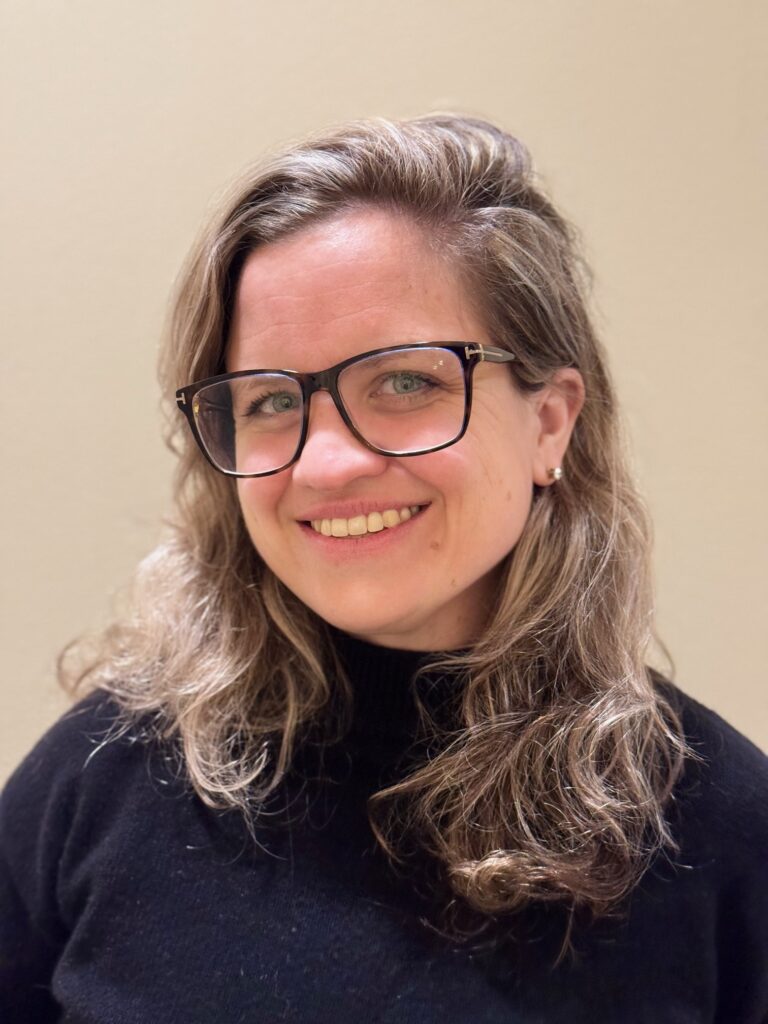 blonde woman wearing a black turtle neck and glasses smiling at camera