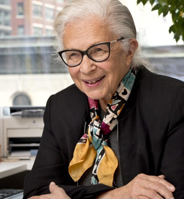 woman in a patterned scarf and black blazer with eyeglasses sits with her arms crossed smiling at the camera