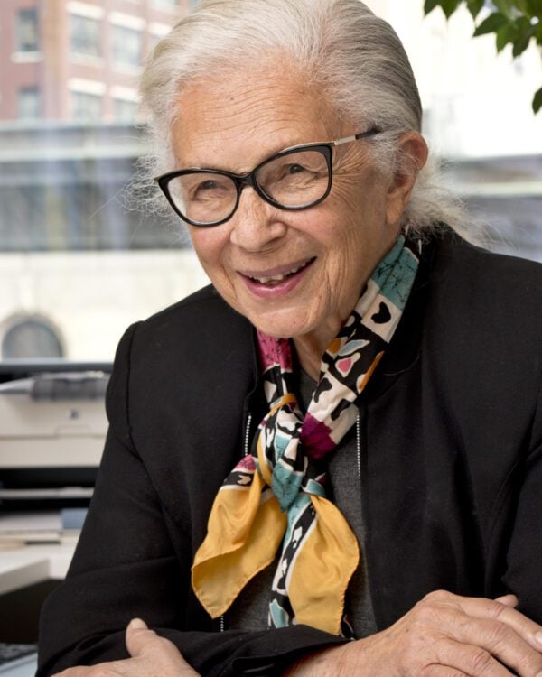 woman in a patterned scarf and black blazer with eyeglasses sits with her arms crossed smiling at the camera