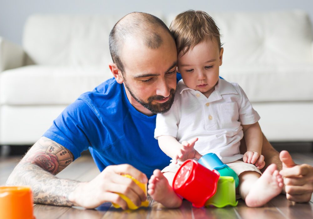 A father plays with his toddler using colorful cups.