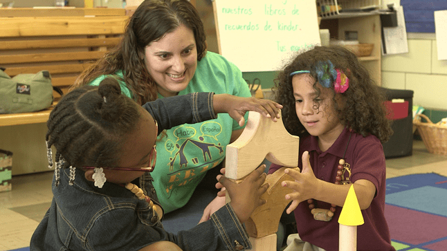 A teacher supervises as two students stack and balance wooden blocks in a classroom.