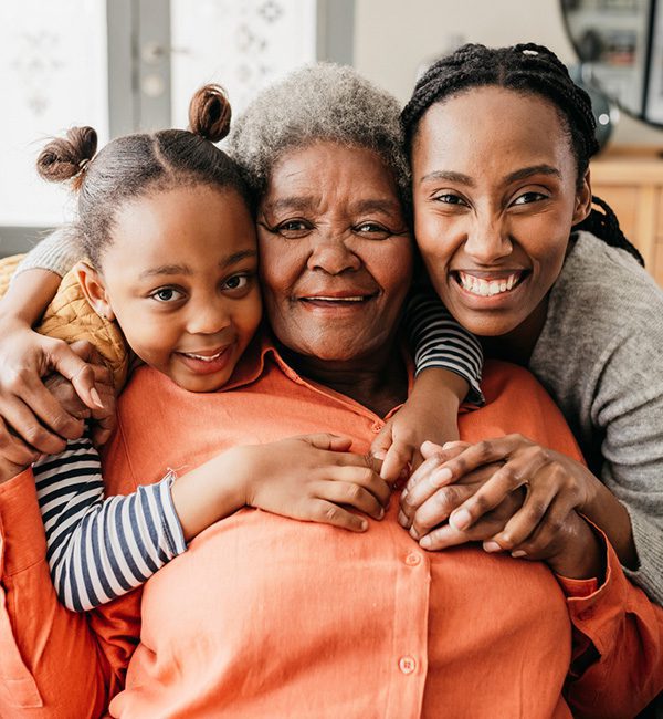 Three generations of a Black family, with a young girl and her mom hugging an older woman in a bright coral sweater from behind while she is seated.