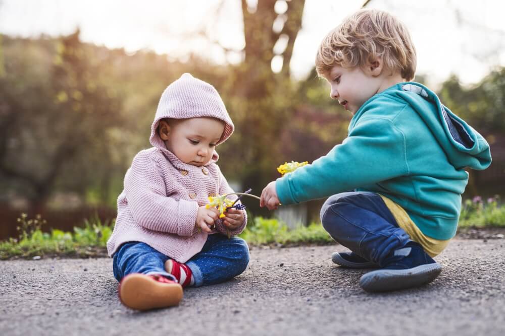 A toddler reaches toward another child on a playground, offering something from her hand